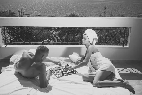 Concetto intelligente e sexy. Attraente coppia giocare a scacchi sulla terrazza nella giornata di sole. Coppia innamorata gioca a scacchi su balcone, natura e mare sullo sfondo. Uomo e signora sexy con asciugamano giocare gioco intellettuale — Foto Stock