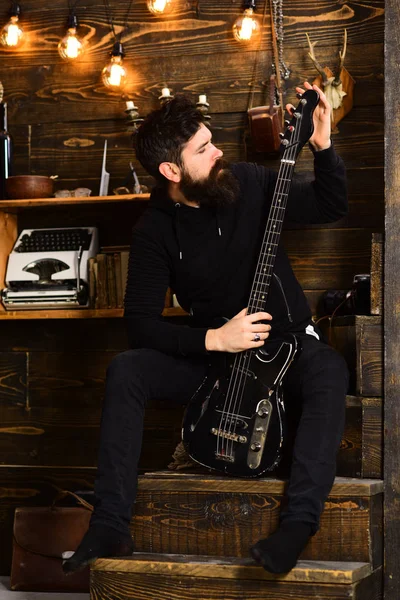 Favourite activity. Man bearded musician enjoy evening with bass guitar, wooden background. Man with beard holds black electric guitar. Guy in cozy warm atmosphere play relaxing soul music