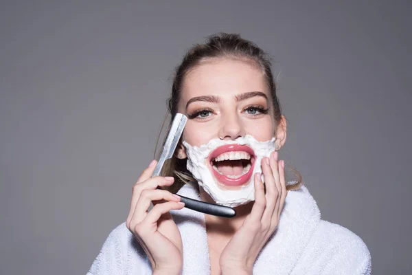 Cheerful blond woman in white bathrobe having fun in bathroom isolated on gray background. Lovely female applying shaving foam on face and holding cut-throat razor, femininity and masculinity concept — Stock Photo, Image