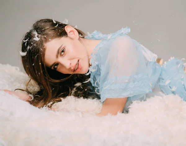 Smiling girl with braces in blue lace nightie lying on soft bed. Cheerful girl with long brunette hair playing with feathers, happy childhood concept