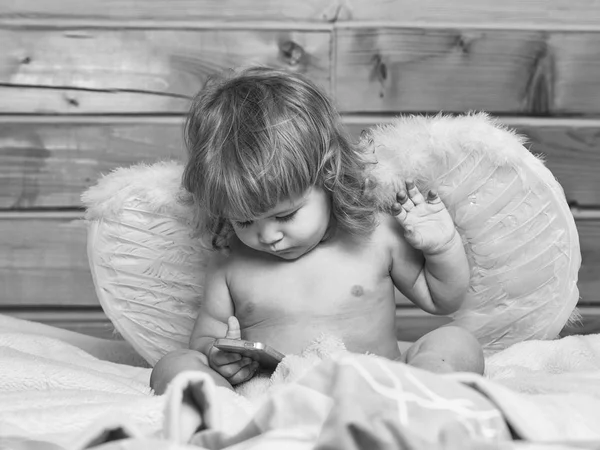 エンジェルは 携帯電話を理解しています 温室風呂白いふわふわタオル裸で座っている濡れている髪のかわいい幸せな美しい遊び心のある子少年羽天使の羽で遊んでの木製の背景上屋内 — ストック写真