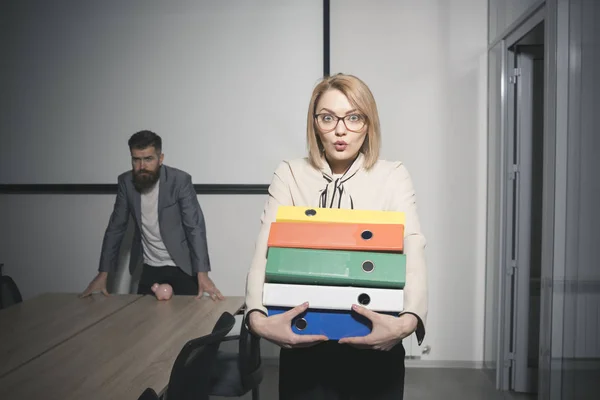 Mujer sorprendida y hombre barbudo de fondo. Mujer de negocios con gafas sostiene carpetas de archivos. Secretaria sexy con documentos de oficina. Concepto de papeleo y burocracia. Día ocupado en la oficina — Foto de Stock