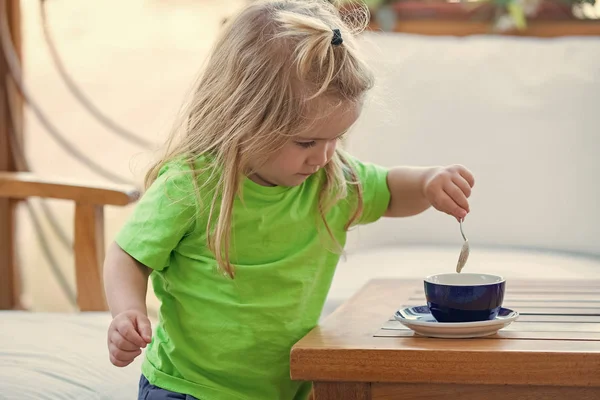 A criança está a fazer café para a mãe. Menino com cabelo longo loiro misturando açúcar com colher de chá . — Fotografia de Stock