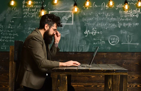 El hombre barbudo trabaja en la computadora portátil en el aula. Barbudo hombre con barba larga con ordenador en pizarra, concepto de nueva tecnología —  Fotos de Stock