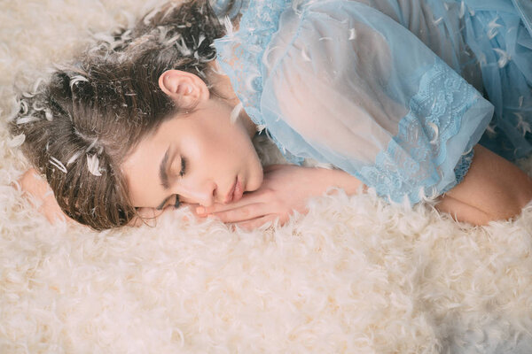 Woman with long hair in tender pajama relaxing. Girl on calm face lay on bed covered with feathers and fluff. Sweet dreams concept. Lady in transparent blue nightie lay on bed and sleep
