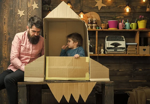 Sueños de volar al espacio. Chico jugar con papá, padre, pequeño cosmonauta sentarse en cohete hecho de caja de cartón. Concepto de lanzamiento de cohetes. Niño lindo chico jugar cosmonauta, astronauta. Niño feliz sentarse en —  Fotos de Stock