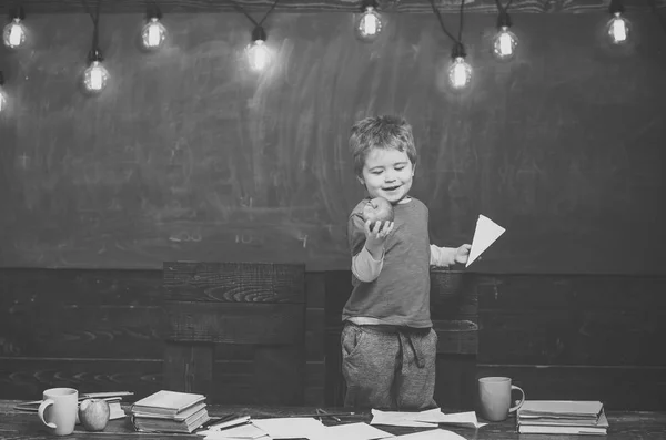 Εναλλακτική εκπαίδευση. Χαμογελαστό αγόρι ψάχνει στο νόστιμο μήλο στο χέρι του. Ξανθό παιδί κρατώντας χαρτί αεροπλάνο. Ευτυχισμένο παιδί στέκεται πίσω από το τραπέζι — Φωτογραφία Αρχείου