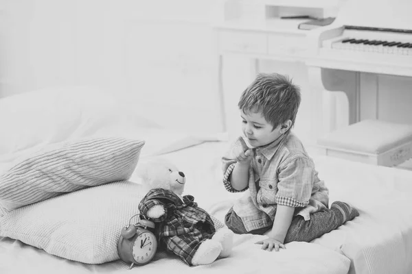 Time to sleep concept. Child in bedroom with silence gesture. Boy with happy face puts favourite toy on bed, time to sleep. Kid put plush bear near pillows and alarm clock, luxury interior background