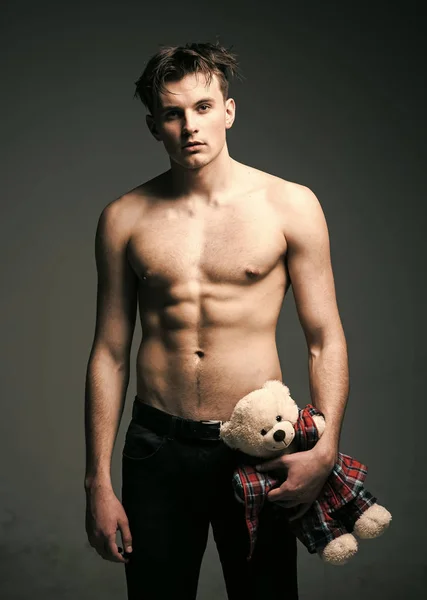 Guy with romantic gift. Man on calm face, muscular figure, holds soft toy plush bear in hand. Man with muscular torso, six packs, looks attractive, dark background. Romantic macho concept
