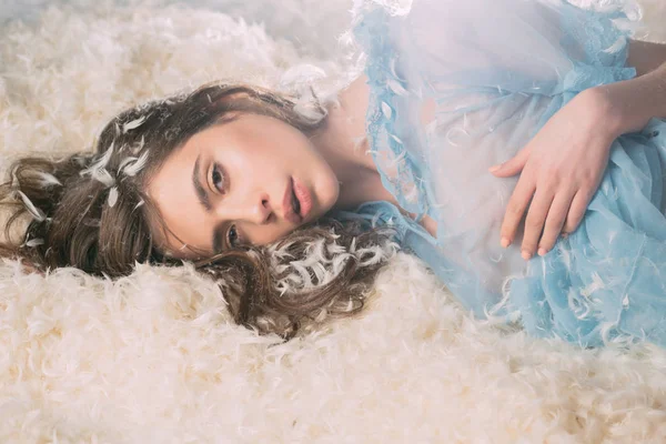 Tender pretty girl enjoying soft embrace of fluffy bed. Beautiful female in blue lace nightgown lying on soft pile of feathers. Young drowsy lady falling asleep, bedtime concept — Stock Photo, Image