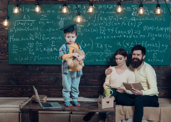 Boy presenting his knowledge to mom and dad. Smart child in graduate cap like to perform. Parents listening their son, chalkboard on background. Support concept. Kid holds teddy bear and performing