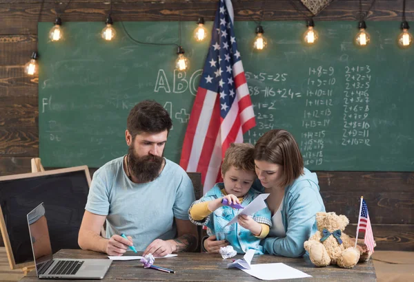 American family at desk with son making paper planes. Homeschooling concept. Parents teaching son to create, handmade. Kid with parents in classroom with usa flag, chalkboard on background — Stock Photo, Image