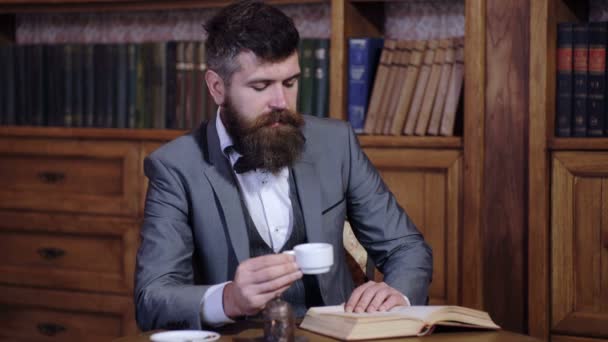 Tyylikäs mies juo teetä ja lukee kirjaa kirjastossa. Mies nauttii kirjan lukemisesta. Parrakas mies luksuspuvussa. Ylellinen elämäntapa, vintage muoti, tee käsite . — kuvapankkivideo