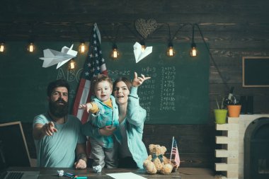 Amerikan ailesinin oğlu ile masada kağıt uçaklar ile oynamak. Evde eğitim kavramı. Ebeveynler sınıf ABD bayrağı ile kara tahta arka plan ile evlat. Anne oğlu Amerikan gelenekleri oynarken öğretim