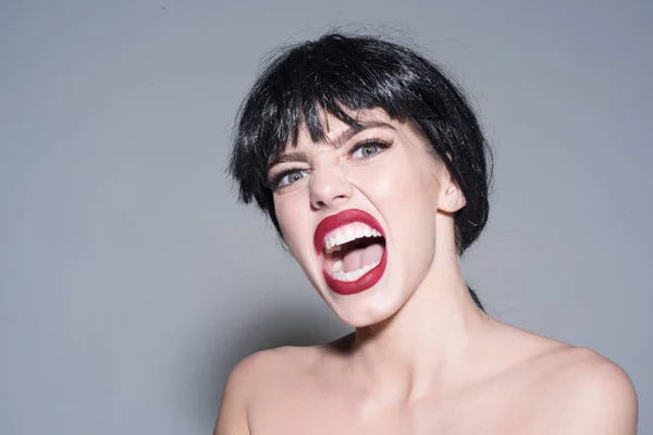 Woman with attractive red lips looks at camera. Lady in black wig with make up on grey background. Scandalous lady concept. Girl on scandalous shouting face posing with naked shoulders — Stock Photo, Image