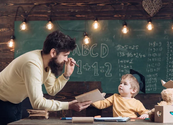 Un concepto prodigioso y genial. Padre, profesor leyendo libro, enseñando niño, hijo, pizarra en el fondo. Papá quiere crecer, hijo genio. Niño con gorra de graduado le gusta escuchar papá — Foto de Stock