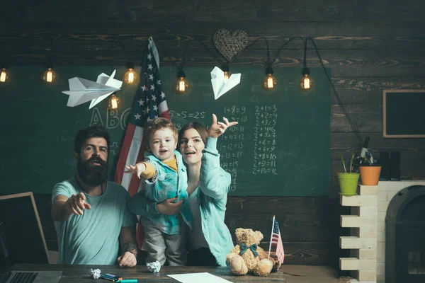 Amerikan ailesinin oğlu ile masada kağıt uçaklar ile oynamak. Evde eğitim kavramı. Ebeveynler sınıf ABD bayrağı ile kara tahta arka plan ile evlat. Anne oğlu Amerikan gelenekleri oynarken öğretim