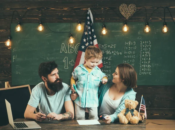 Amerykańska rodzina siedzieć przy biurku z flagą syn i usa. Dziecko z rodzicami w klasie z flagą usa, tablica na tle. Koncepcja edukacji patriotycznej. Rodziców, uczy syna amerykańskiej tradycji gry — Zdjęcie stockowe