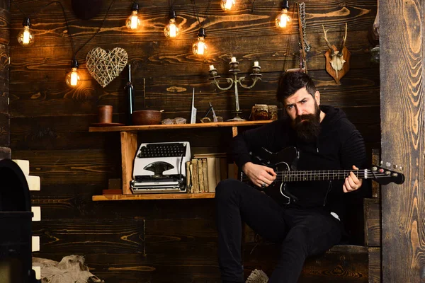 Betrokken bij ontspanning. Man met baard heeft zwarte elektrische gitaar. Guy in gezellige warme sfeer spelen ontspannende muziek. Man met baard musicus geniet van avond met basgitaar, houten achtergrond — Stockfoto