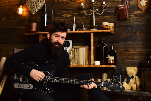 Favoriete activiteit. Man met baard musicus geniet van avond met basgitaar, houten achtergrond. Guy in gezellige warme sfeer spelen ontspannende soul muziek. Man met baard houdt zwarte elektrische gitaar — Stockfoto