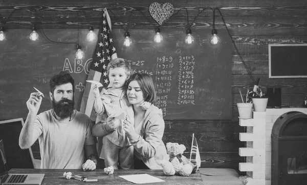 Módní doplňky. Americká rodina u stolu s synem, takže papír letadla. Koncepce knihy. Dítě s rodiči v učebně s vlajkou usa, tabuli na pozadí. — Stock fotografie