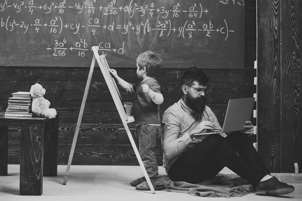 math lesson. Arithmetic lesson at school. Kid writing on chalkboard while concentrated teacher works on laptop. Side view sitting man and standing kid back to back