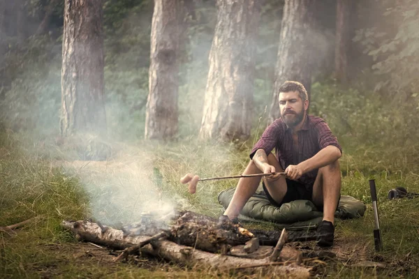 Лагерь отдыха. Человек путешественник жареные колбасы на палочке на костре в лесу — стоковое фото