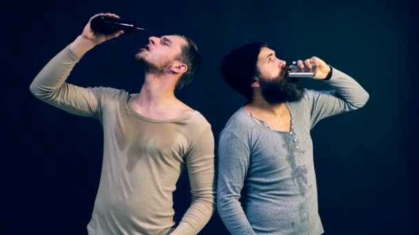 Dois homens bebem de garrafas em um fundo preto. O problema do alcoolismo. A dependência do álcool é um problema da sociedade moderna . — Vídeo de Stock