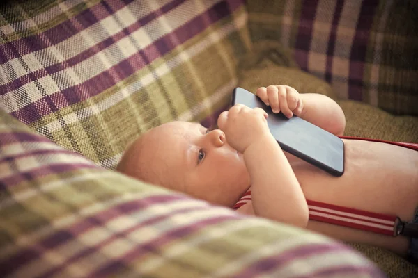 child is studying the phone. Baby boy with suspenders lie in armchair