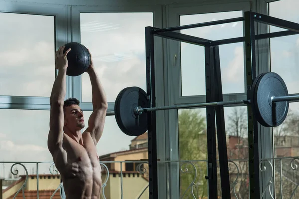 Sport- und Fitnesskonzept. Sportler, Athlet, muskulöser Macho trainiert mit Medizinball, Fenster und Langhantel im Hintergrund. Mann mit nacktem Oberkörper im Fitnessstudio genießen Training, trx — Stockfoto