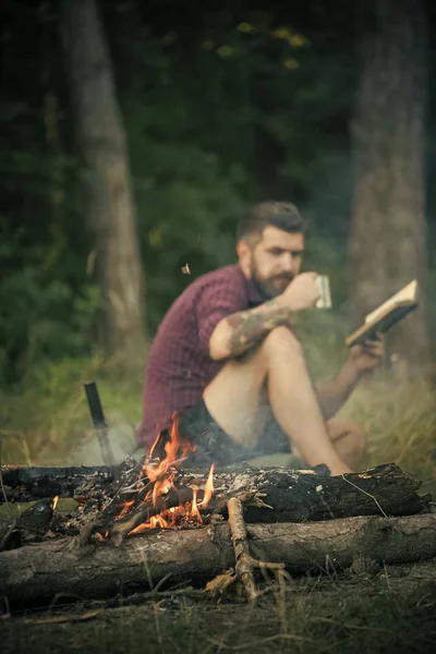 outdoor recreation. Hipster hiker with book and mug at bonfire in forest