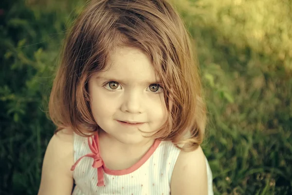 A bájos kislány. Lány, barna szemek, az aranyos arca ül a fűben — Stock Fotó