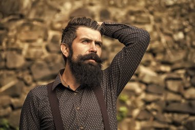 Erkek vahiy. Siyah saçlı ve mavi gömlek ve mor pantalon taş duvar gri arka planda, yatay resim açık duran uzun gür sakallı bir üst düzey yakışıklı closeup görünümü