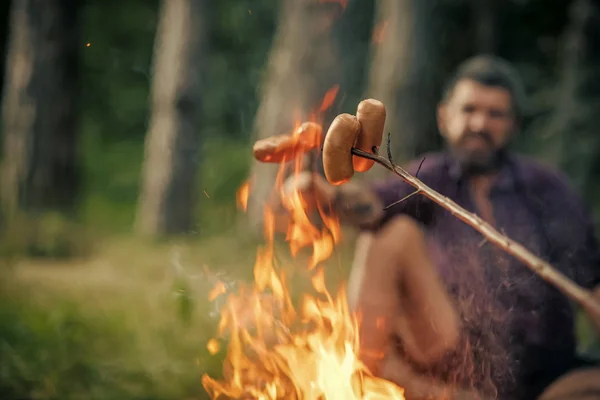 Camping brand. Worstjes geroosterd op vreugdevuur met wazig man op natuurlijke achtergrond — Stockfoto