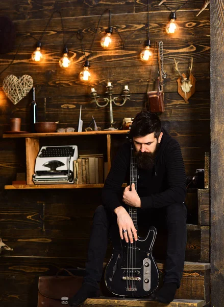 Connection through music. Man bearded musician enjoy evening with bass guitar, wooden background. Man with beard holds black electric guitar. Guy sits thoughtful dreamy in cozy warm atmosphere — Stock Photo, Image