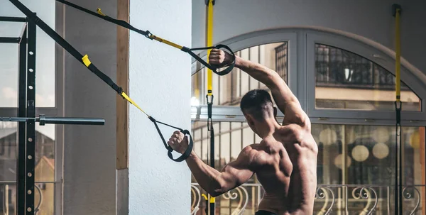 Sportsman, atleta, macho muscolare fa esercizio con loop trx, finestra sullo sfondo. Uomo con busto nudo e schiena muscolosa in palestra godere di allenamento, trx, vista posteriore. Sport e concetto di palestra — Foto Stock