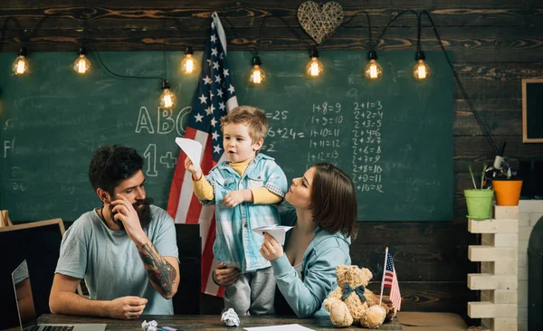 Parents enseignant fils traditions américaines jouer. Enfant avec parents en classe avec drapeau américain, tableau en arrière-plan. Famille américaine au bureau avec fils fabriquant des avions en papier. Concept d'enseignement à domicile — Photo