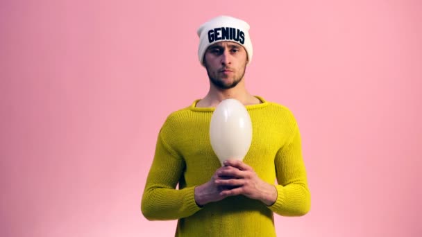 Der Typ im gelben Pullover mit weißem Hut auf rosa Hintergrund streckt eine große Lampe in seinen Händen. Geniales Konzept. — Stockvideo