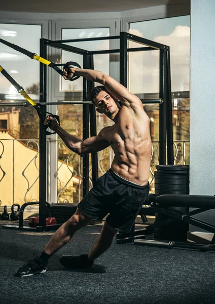 Conceito de desporto e ginásio. Homem com tronco nu, e peito muscular no ginásio desfrutar de treinamento, trx. Esportista, atleta, musculoso macho faz exercício com loops trx, janela no fundo — Fotografia de Stock