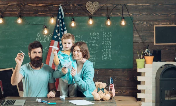 American family at desk with son making paper planes. Homeschooling concept. Kid with parents in classroom with usa flag, chalkboard on background. Parents teaching son american traditions playing — Stock Photo, Image