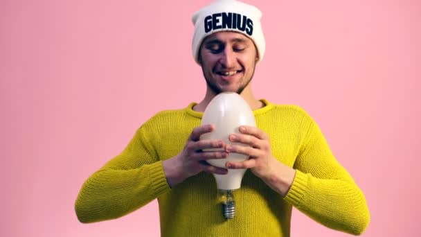 Jonge man in een gele trui glimlachend schudt een grote lamp op een roze achtergrond. Idee concept. — Stockvideo