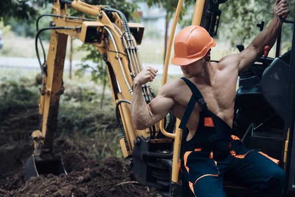 Employee concept. Employee flex biceps and triceps muscle. Strong employee at excavator cabin. Employee in working uniform on excavation site. Strength and masculinity