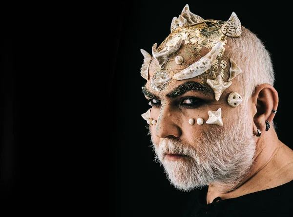 Fancy Halloween make-up, actor portraying ugly monster with thorns on face. Side view bearded man with evil eyes wearing silver ear rings isolated on black background