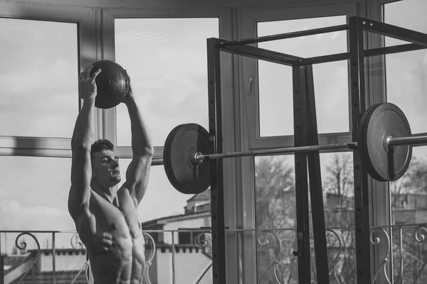 Concetto di sport e palestra. Sportivo, atleta, macho muscolare fa esercizio con palla medica, finestra e bilanciere sullo sfondo. Uomo con torso nudo in palestra godere di formazione — Foto Stock