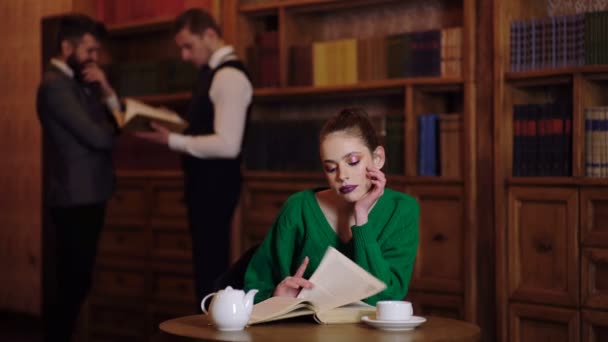 ライブラリの概念。少女は本を反転し、ライブラリで、二人の男が話す背景で茶を飲む. — ストック動画