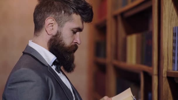 De bebaarde man leest een boek staan op de boekenplank in de bibliotheek. Concept van het lezen van boeken. — Stockvideo