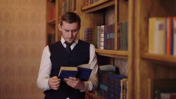 Een man in een pak leest een groot boek staande in de buurt van een boekenkast in een bibliotheek. Leren concept. — Stockvideo