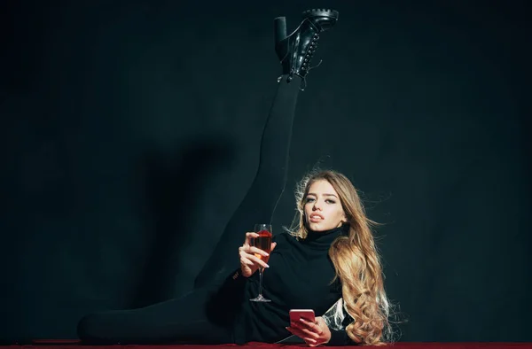 Fashionabla flexibla dam med make up och smal benet låg, svart bakgrund. Tjej med långt hår höjer benet, innehar glas vin och smartphone, kopia utrymme. Mode och skönhet koncept. — Stockfoto
