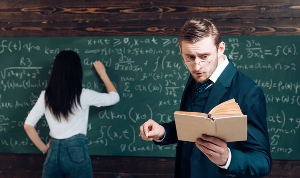 Doing sums. Rear view woman write equation on chalkboard. Learning math enables me to think clearly. Teacher man in glasses read problem statement from textbook to girl student. School day