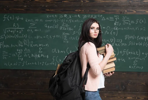 Inteligente y seguro. Estudiante en gafas con pila de libros y soporte de mochila en pizarra en el aula. La literatura me interesa. Educación en casa. Gusano de biblioteca. Me encanta leer. — Foto de Stock